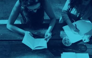 Two women sat at park bench reading and writing