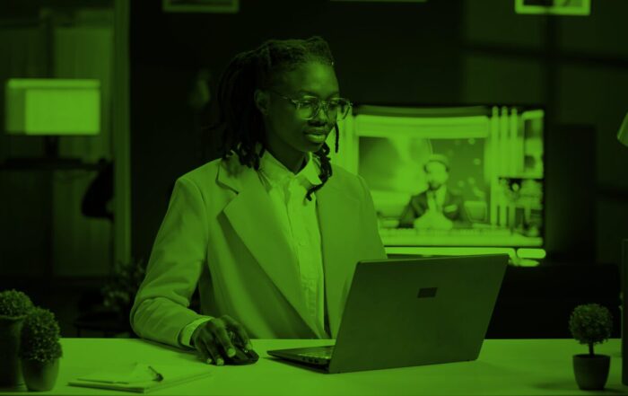 A woman wearing a blazer sits working on her laptop. Behind her is a TV screen showing a news broadcast.