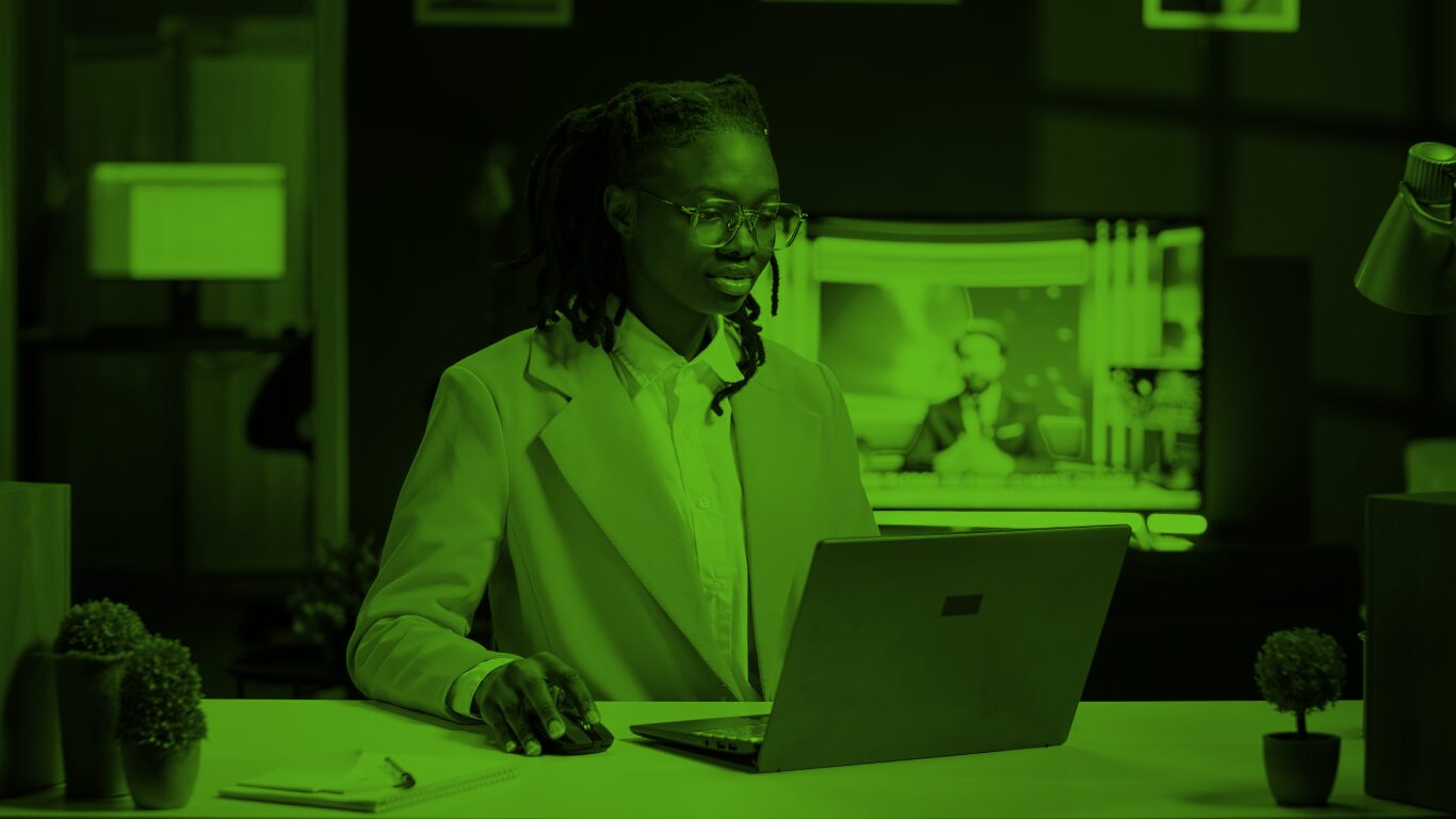 A woman wearing a blazer sits working on her laptop. Behind her is a TV screen showing a news broadcast.