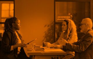 A woman speaks to a woman and man sitting across a desk from her in a workplace.