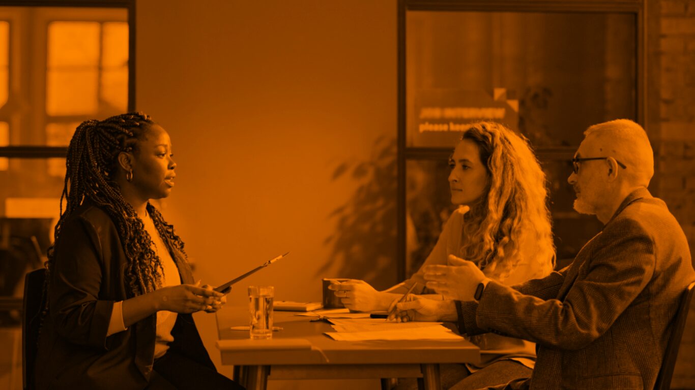 A woman speaks to a woman and man sitting across a desk from her in a workplace.