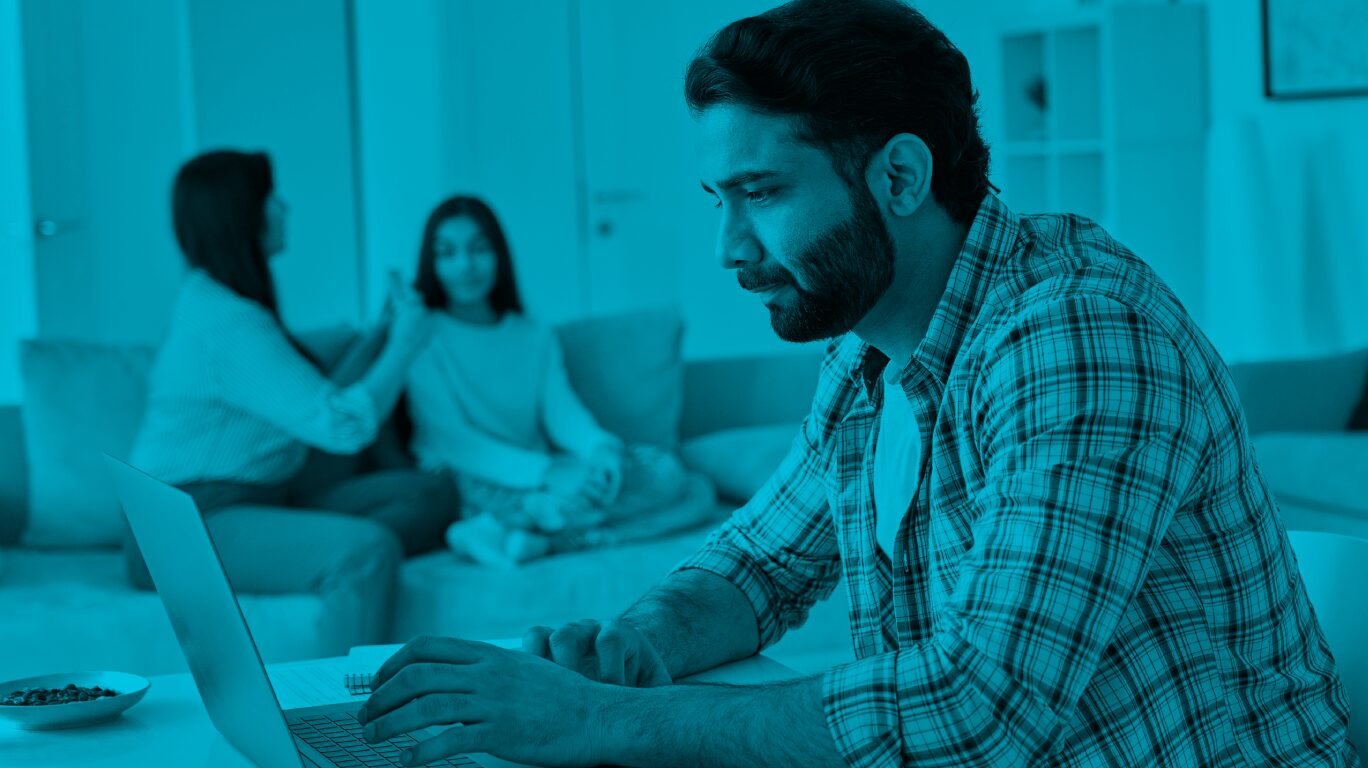 A man types on his laptop while a woman and child talk in the background.