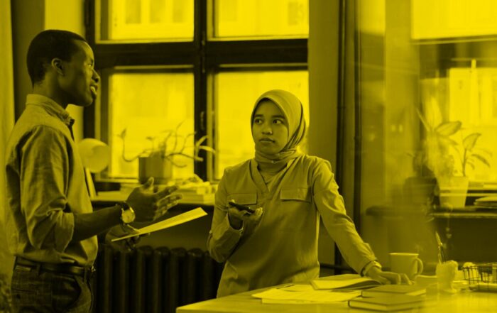 A woman in an office stands with her phone in her hand listening to a male colleague talk to her.