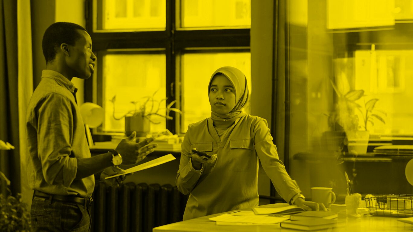 A woman in an office stands with her phone in her hand listening to a male colleague talk to her.