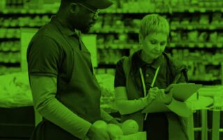 A woman working in a grocery store takes notes while talking to a man holding a box of fruit.