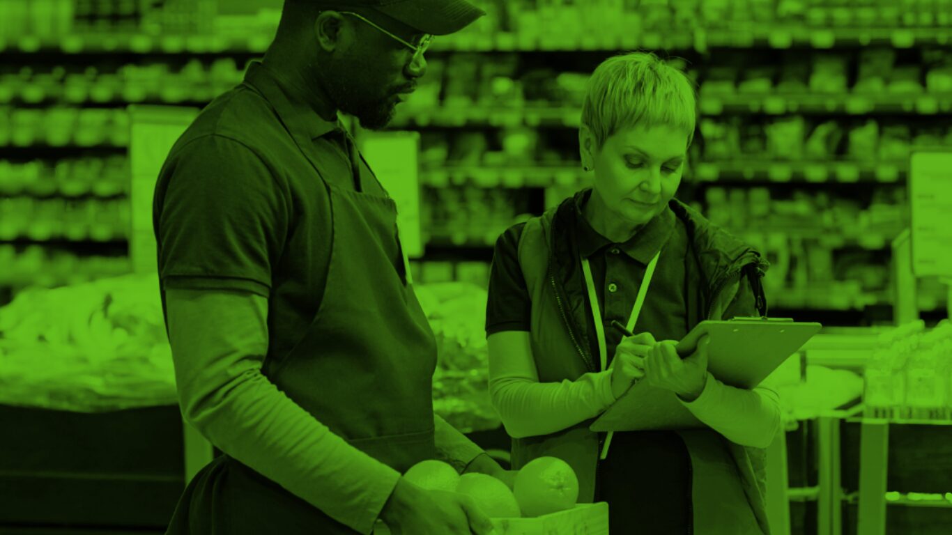 A woman working in a grocery store takes notes while talking to a man holding a box of fruit.