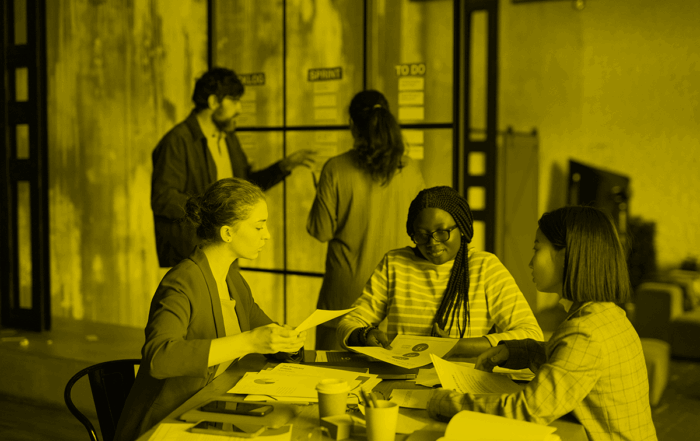 Three women of different races talk at a table in an office. There is a man talking to a woman in the background.