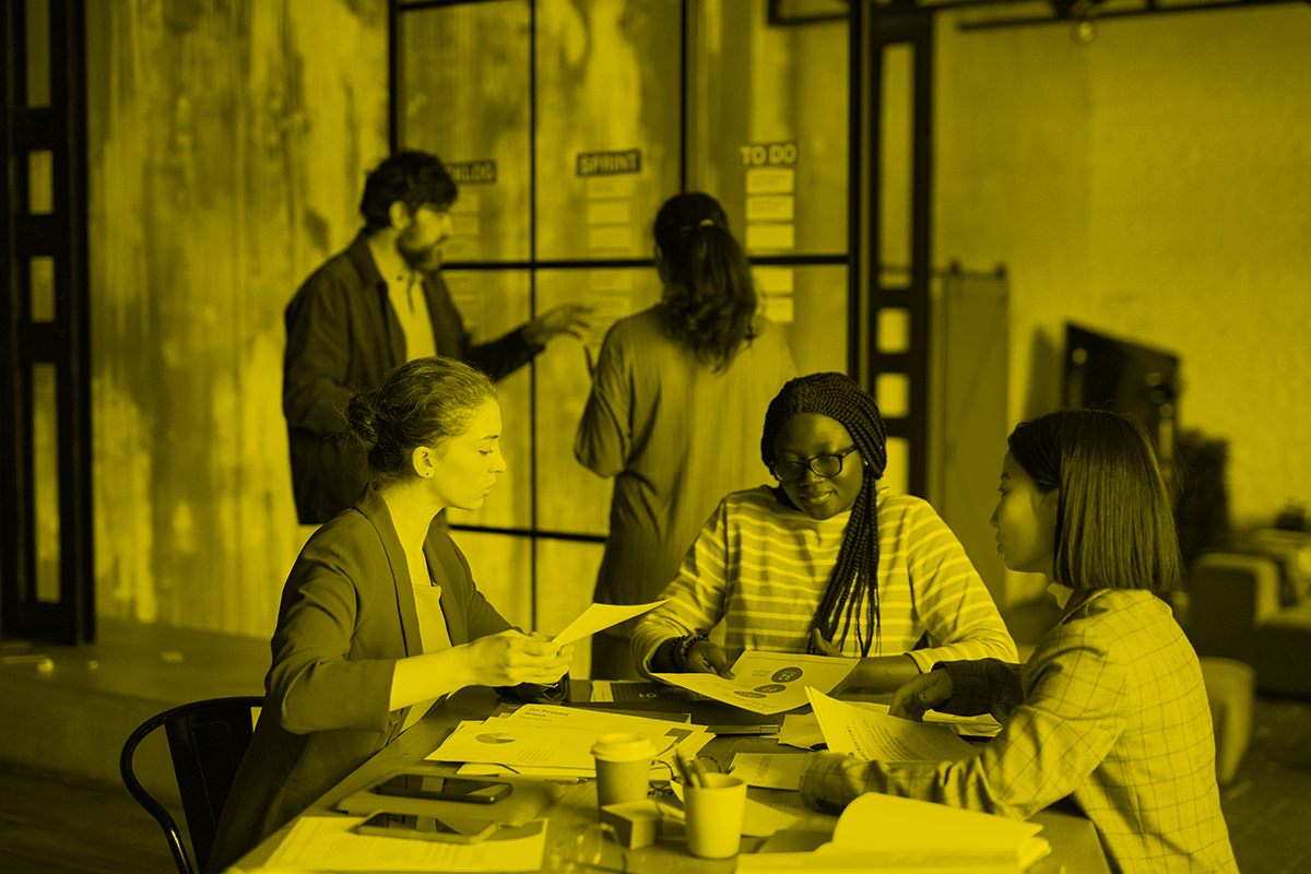 Three women of different races talk at a table in an office. There is a man talking to a woman in the background.